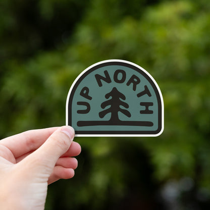 Fingers hold the Up North sticker against a soft green wall of out of focus foliage.