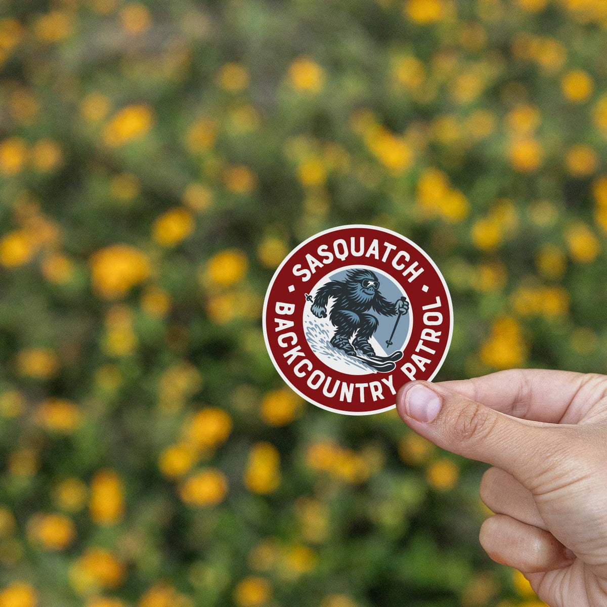 Yellow blooms blur behind fingertips holding the Sasquatch Backcountry Patrol sticker steady.