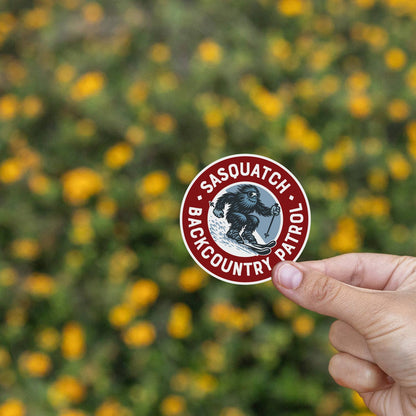 Yellow blooms blur behind fingertips holding the Sasquatch Backcountry Patrol sticker steady.