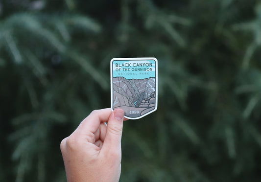 Held further back against the same dark pine boughs, the Black Canyon of the Gunnison sticker shows its 1999 date.