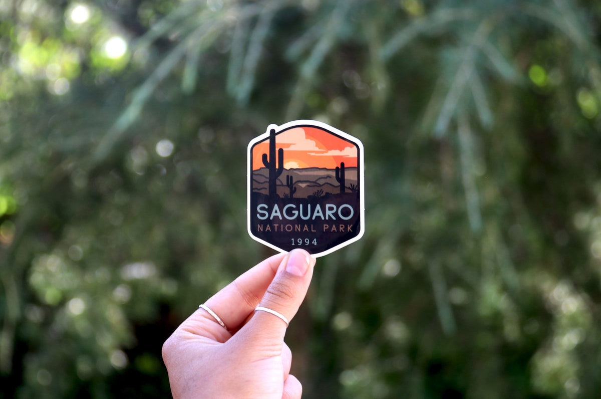 Held farther back against blurred green branches, the Saguaro National Park sticker shows its 1994 badge shape.