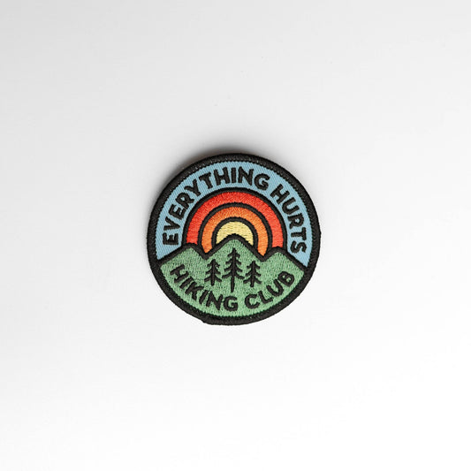 Round Everything Hurts Hiking Club patch rests on plain white, its rainbow arching over green hills and pines.