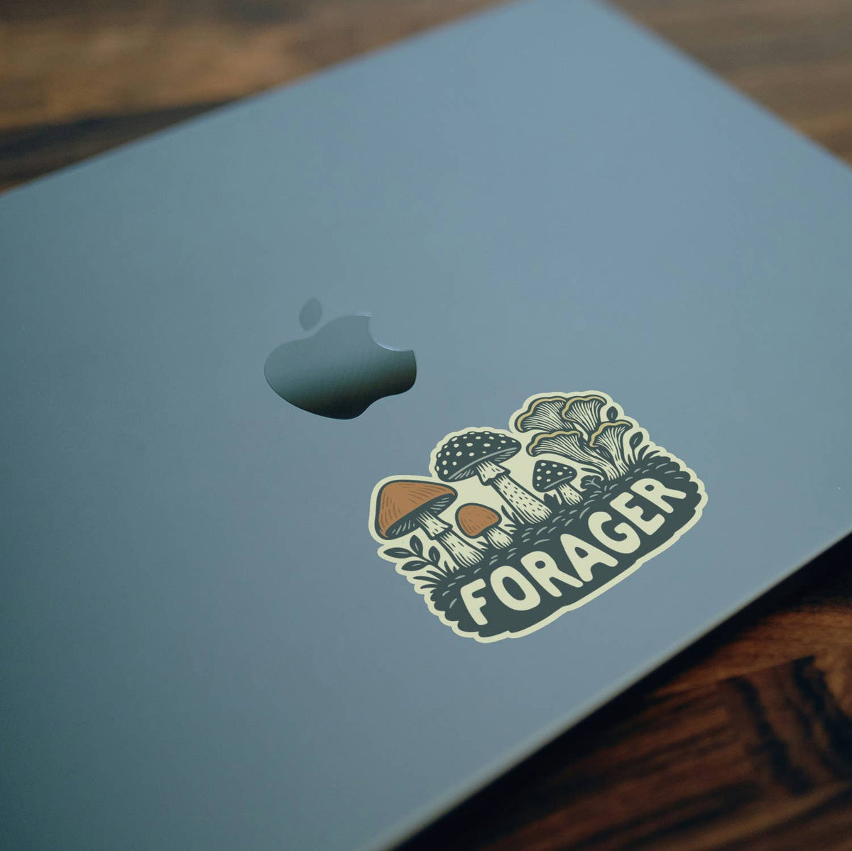 Stuck near the corner of a grey laptop lid, the Forager Mushrooms sticker sits on a wooden desk.