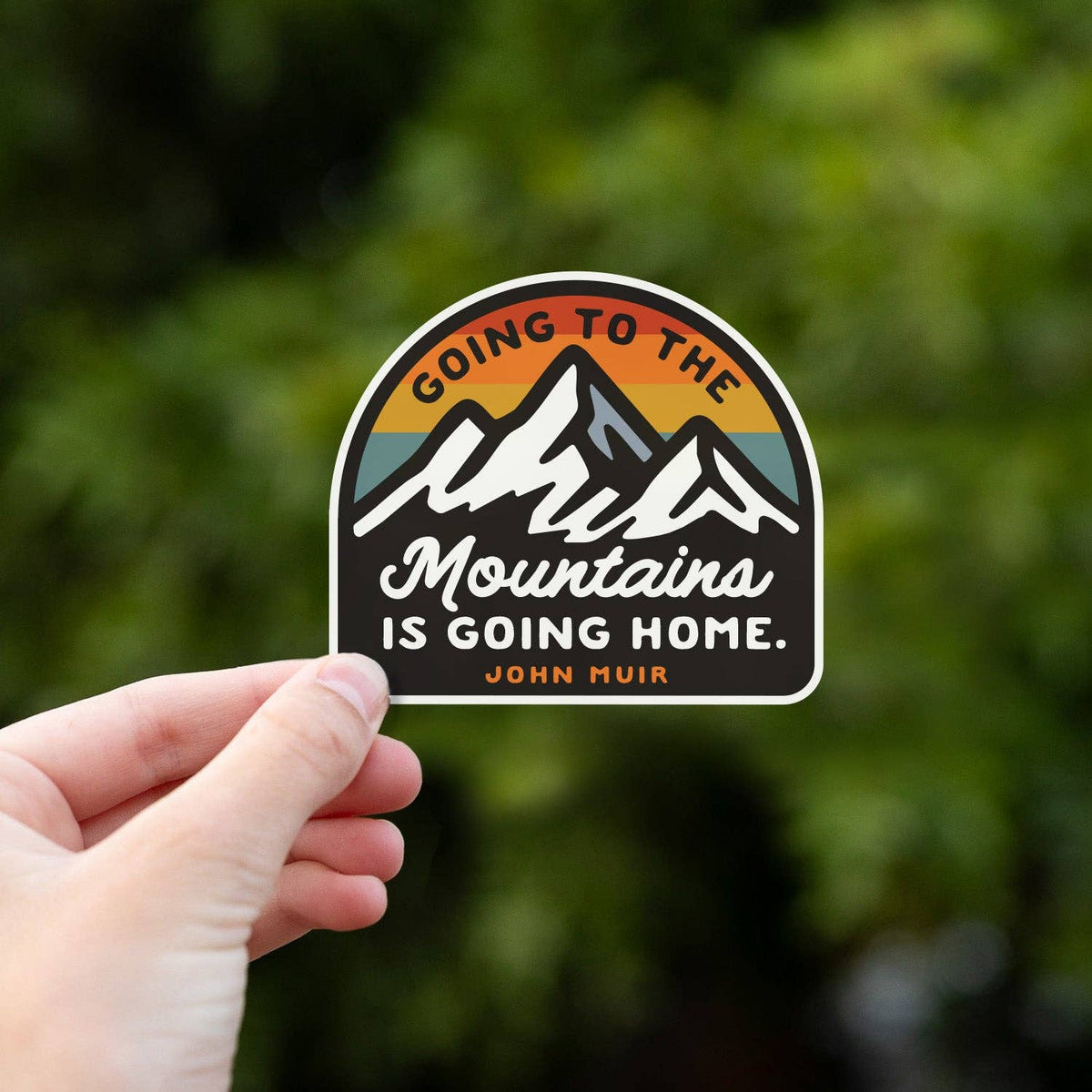 A hand pinches the Going to the Mountains is Going Home sticker in front of blurred green leaves.