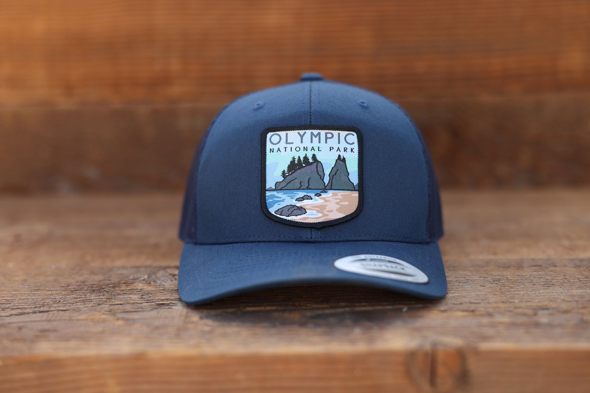 Set on weathered planks, the navy Olympic National Park trucker hat faces forward with brim sticker in place.