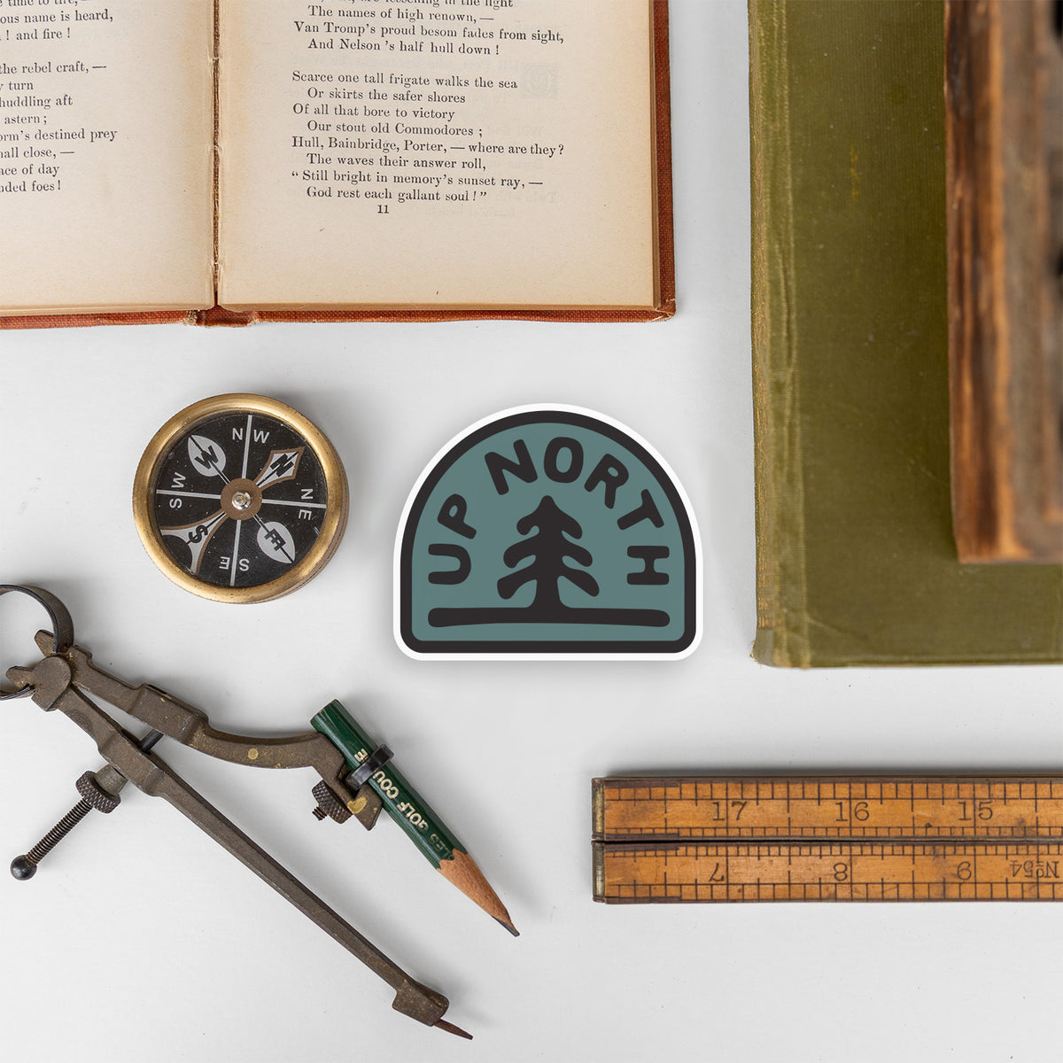 The Up North sticker lies on a desk among an open poetry book, a brass compass, and a wooden ruler.