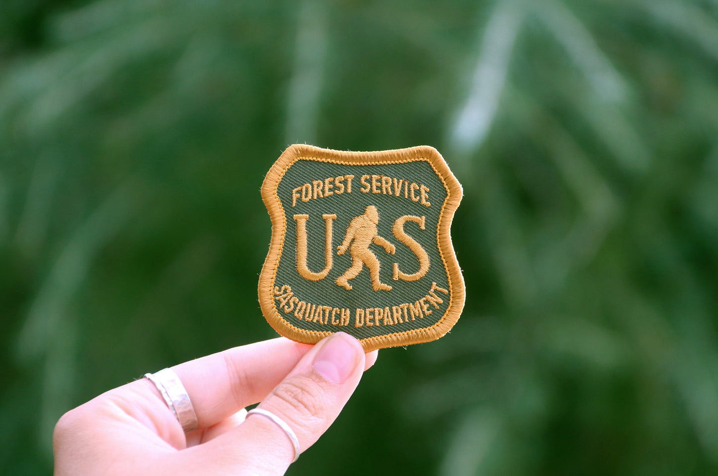 Fingers pinch the US Sasquatch Department patch in front of soft green needles outdoors.