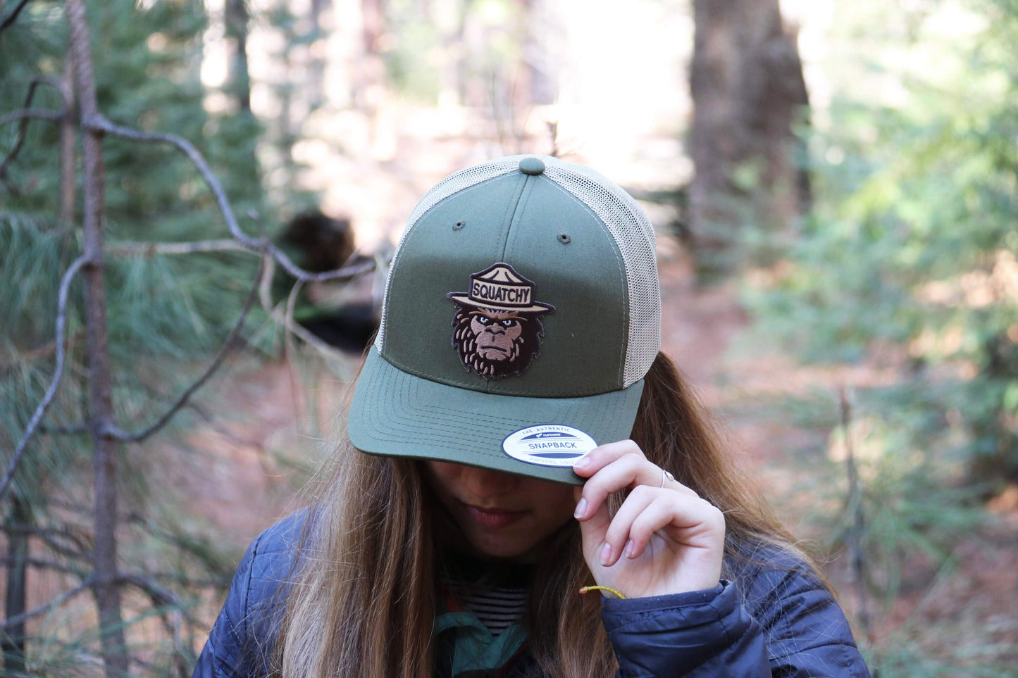 Tugging the brim of a green trucker hat with the Squatchy patch on front, a woman stands among pines.