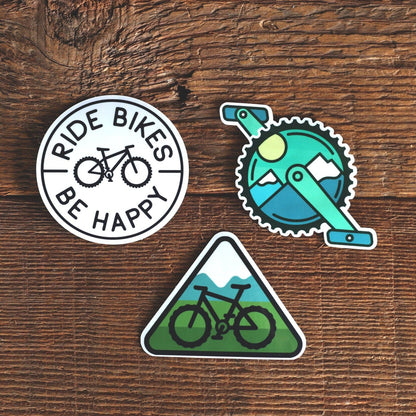 Three Mountain Bike Sticker Set decals rest on dark wood, showing Ride Bikes Be Happy, a crank and a trail sign.