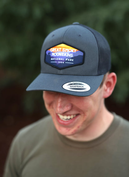 A smiling man looks down in the charcoal Great Smoky Mountains trucker hat with evergreens blurred behind him.
