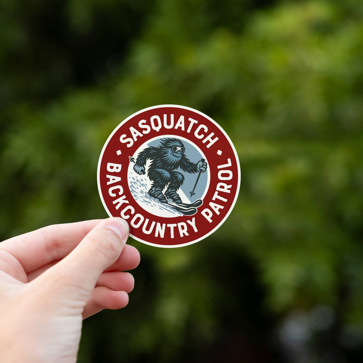 Close up in a hand, the Sasquatch Backcountry Patrol sticker fills the frame with leafy green behind it.