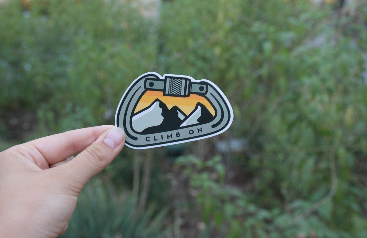 Held in front of leafy green brush, the Climb On Carabiner sticker shows its orange sunset and peaks.