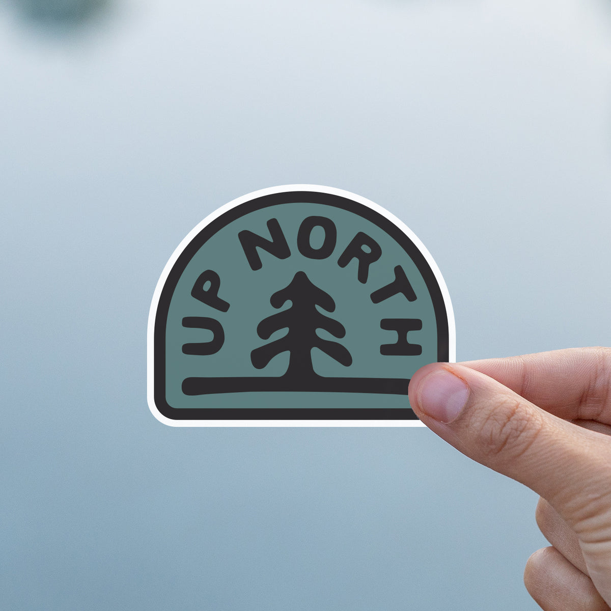 A thumb and finger hold the Up North sticker against a pale grey lake backdrop.