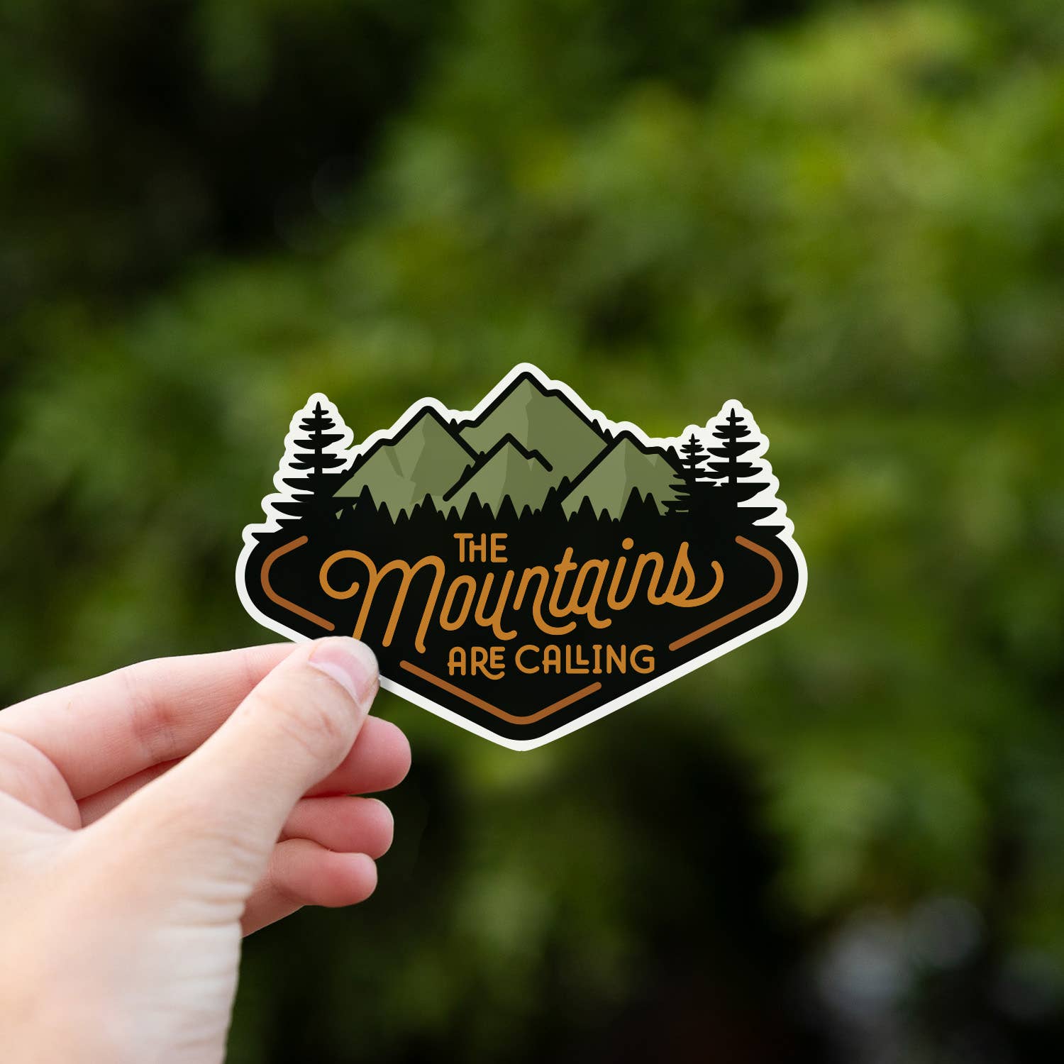 Held up to leafy summer green, The Mountains Are Calling sticker shows its orange outlined border.