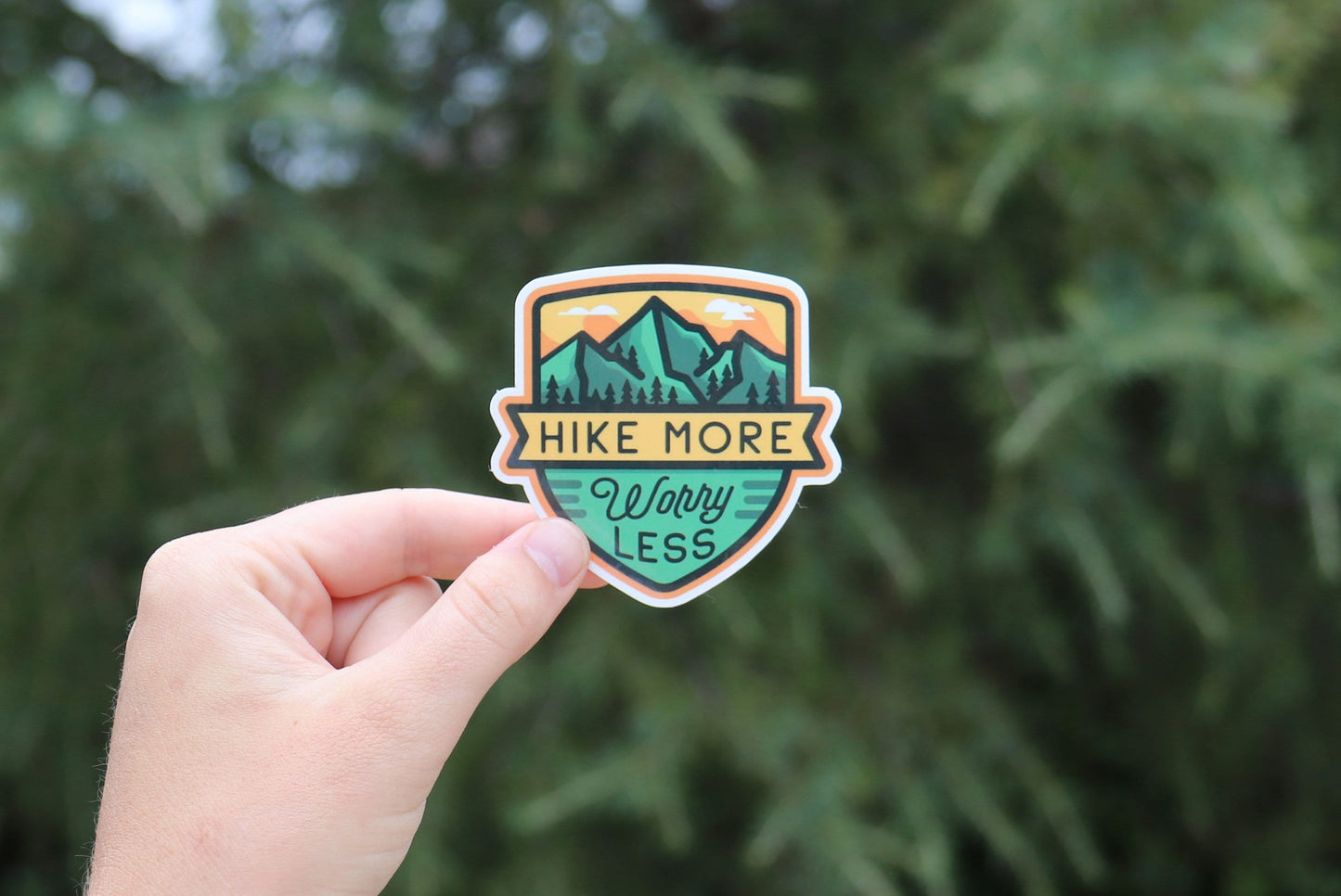 In front of a thick evergreen bough, the Hike More Worry Less sticker shows its teal peaks and script.
