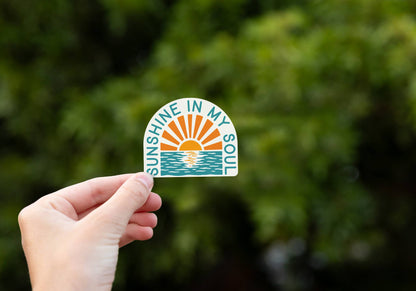 A hand holds the Sunshine In My Soul sticker, its orange sun and teal water bright against blurred green leaves.
