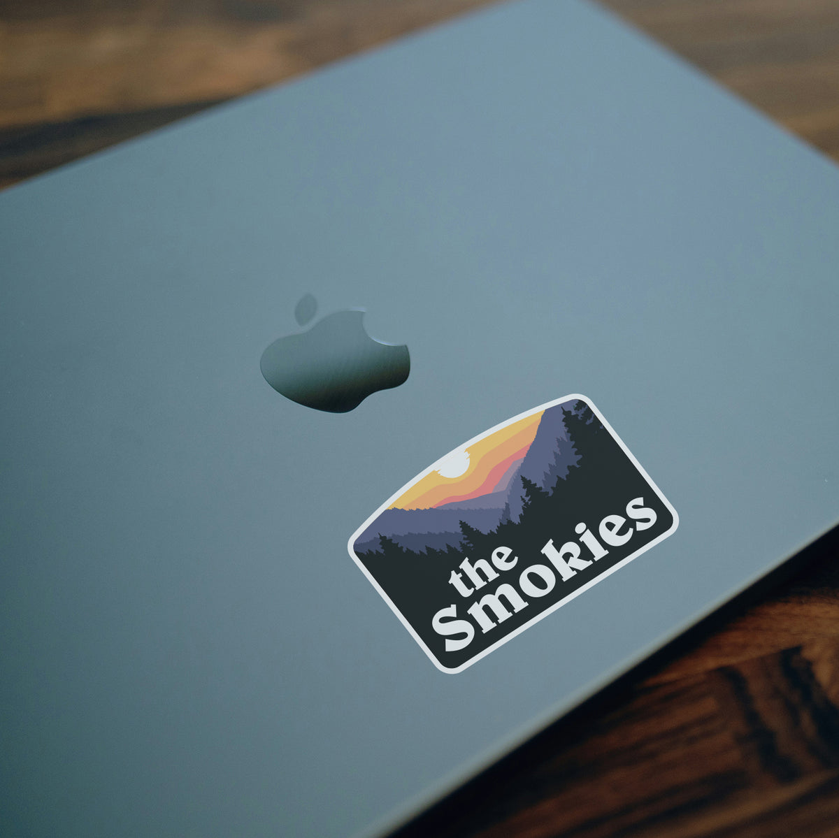 On a closed laptop lid, the Smokies sticker sits beside the dim Apple logo near a wooden desk edge.