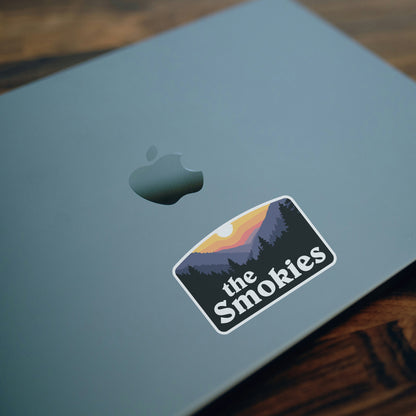 On a closed laptop lid, the Smokies sticker sits beside the dim Apple logo near a wooden desk edge.
