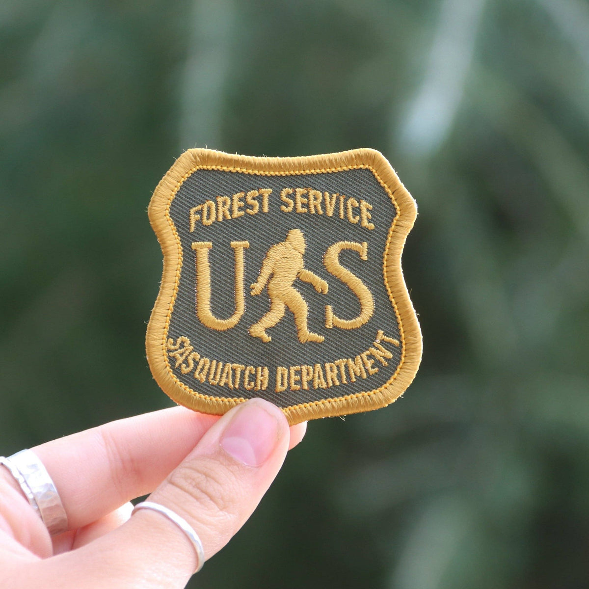 A ringed hand holds the US Sasquatch Department patch up against blurred green foliage.