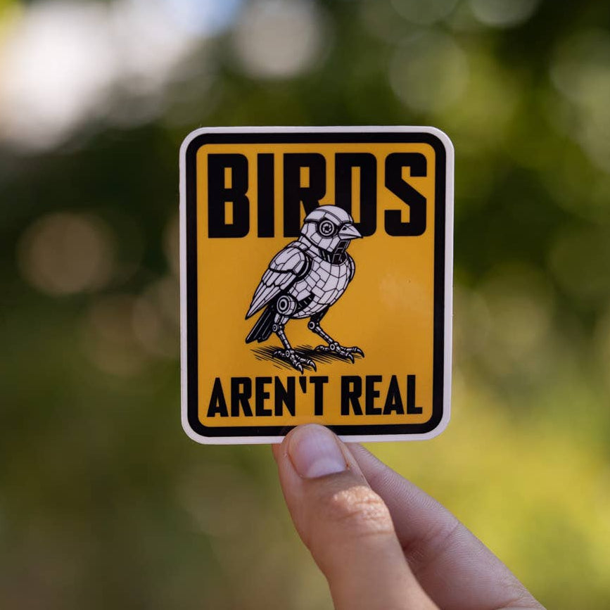 A hand holds up the yellow Birds Aren't Real sticker with its perched crow, green leaves blurred behind.