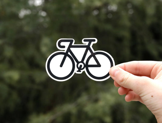 Fingers hold the black and white Road Bike sticker against blurred evergreen branches in flat daylight.