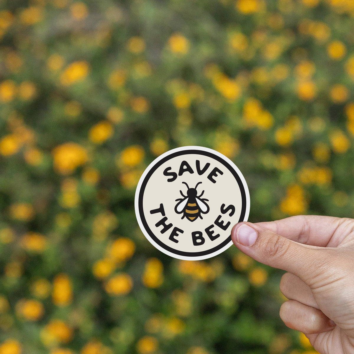 A hand pinches the Save The Bees sticker above a blurred field of small yellow wildflowers.