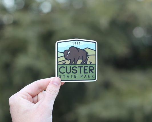 Backed by soft daylight through the trees, the Custer State Park sticker shows the 1912 date above the bison.