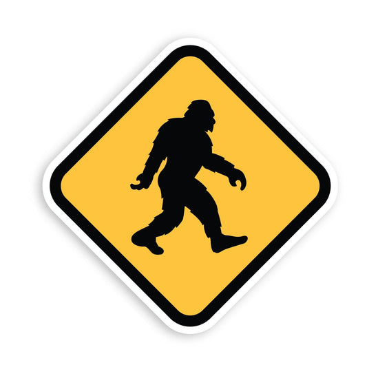 The diamond shaped Bigfoot Crossing sticker sits on white, its black sasquatch striding across a yellow field.