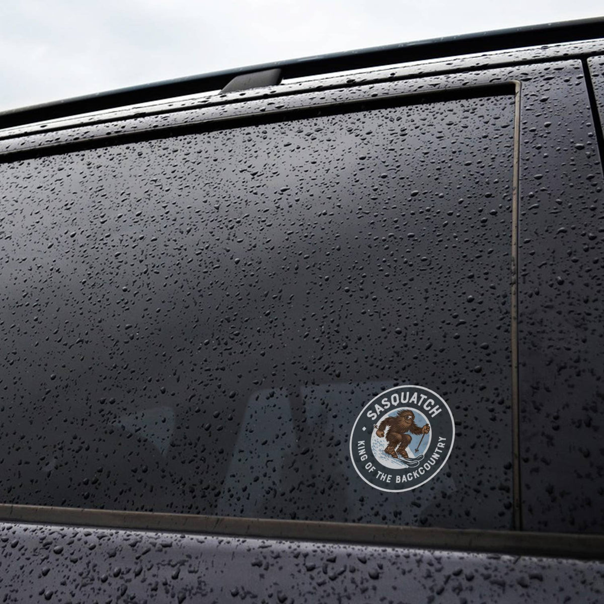 Stuck to the rain beaded window of a dark car, the small Sasquatch King of the Backcountry sticker catches light.