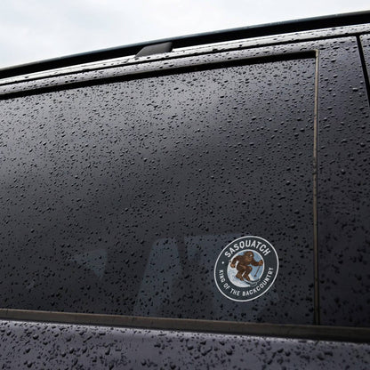 Stuck to the rain beaded window of a dark car, the small Sasquatch King of the Backcountry sticker catches light.