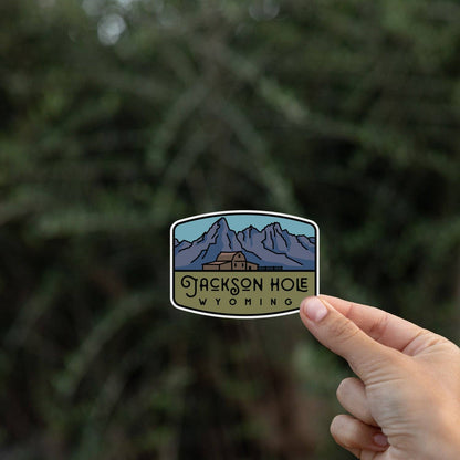 Against dark evergreen branches, a hand steadies the Jackson Hole Wyoming sticker by its lower corner.