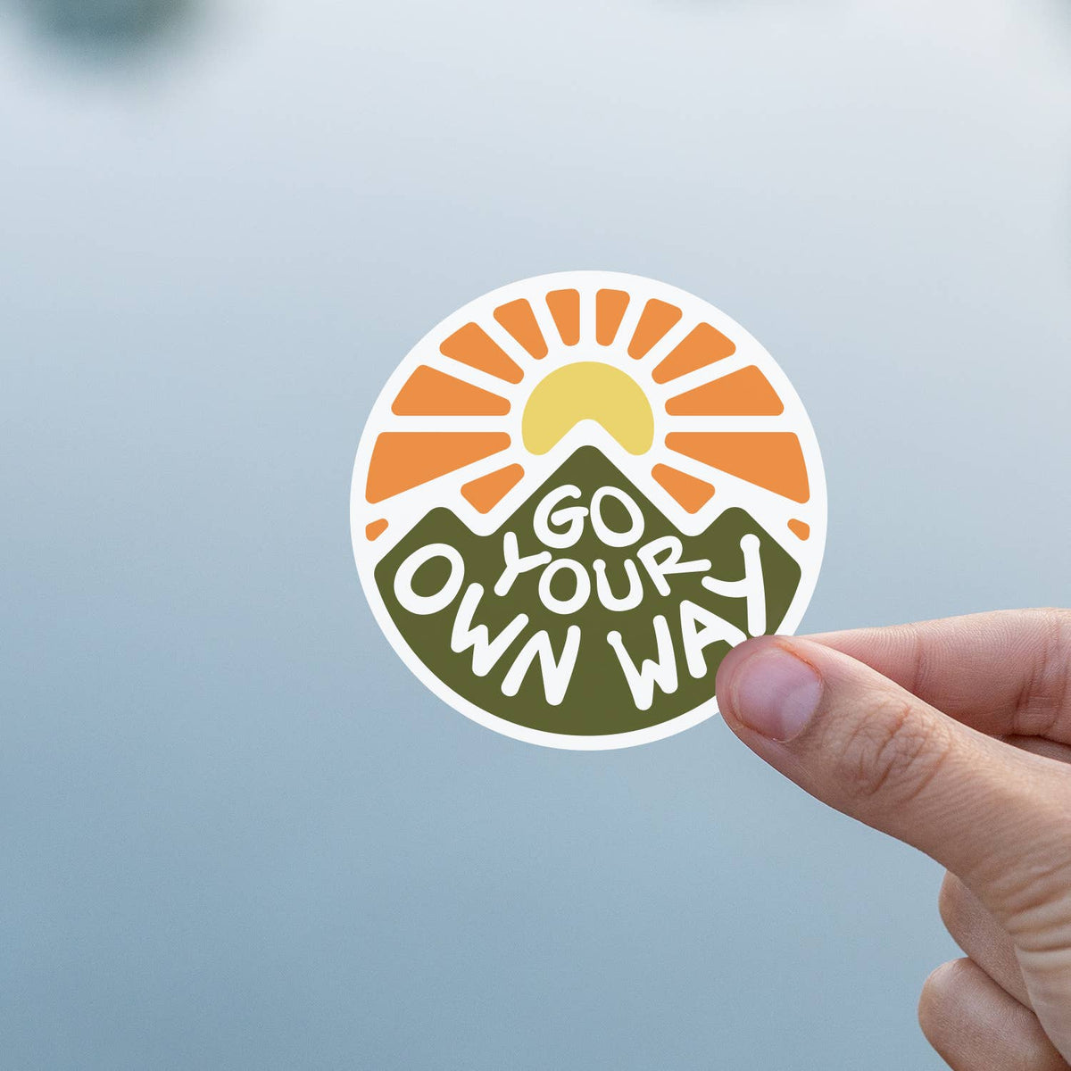 Against a soft blue grey background, a thumb and finger hold the Go Your Own Way sticker.