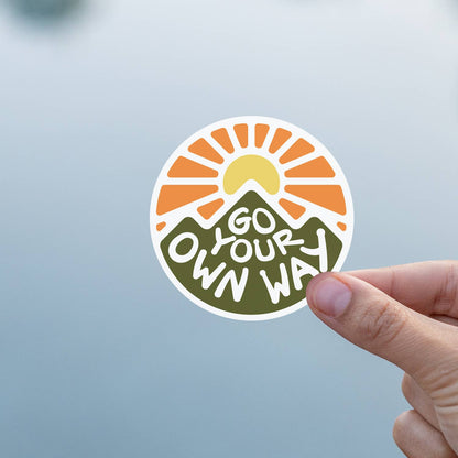 Against a soft blue grey background, a thumb and finger hold the Go Your Own Way sticker.