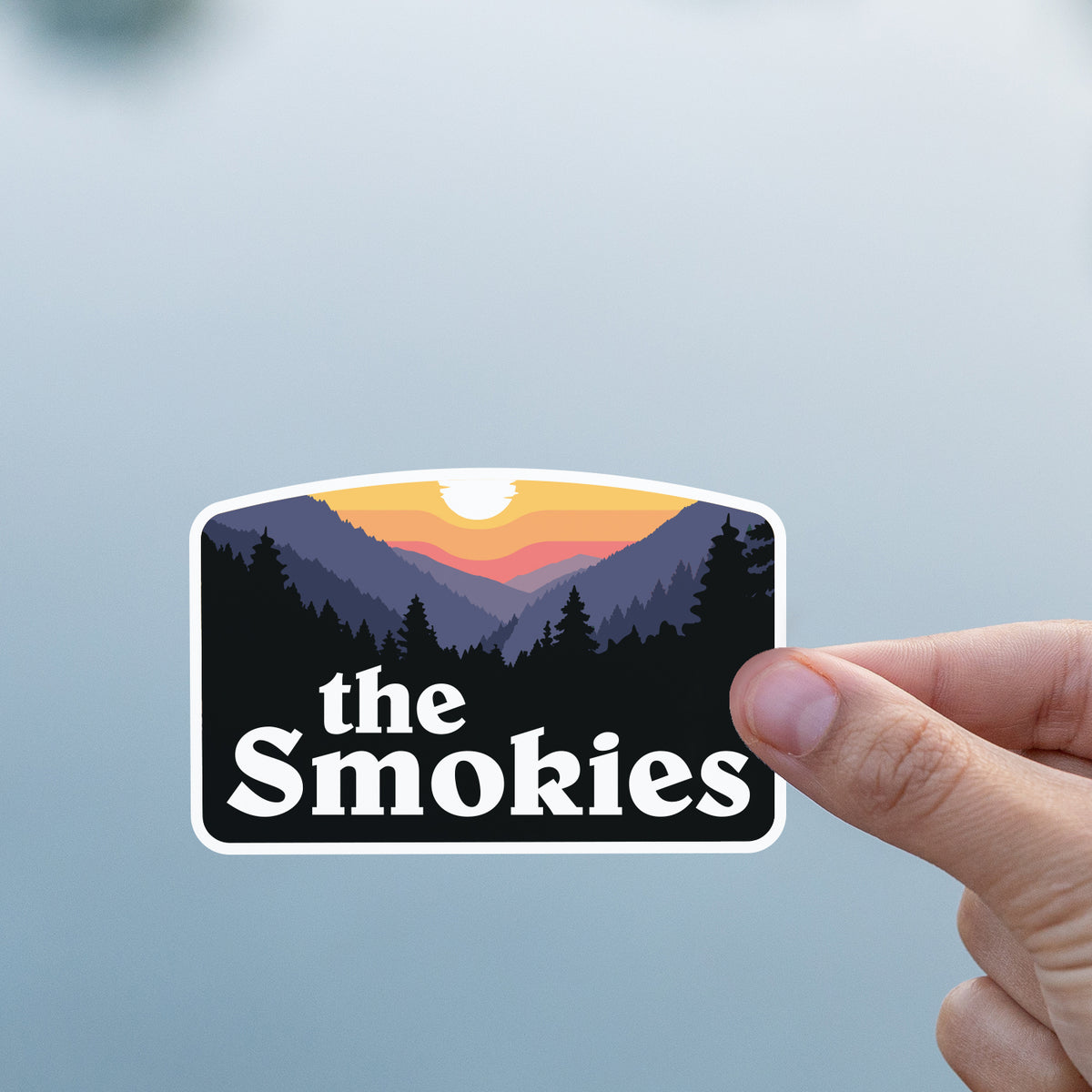 A thumb and finger pinch the The Smokies sticker against a smooth grey water surface.