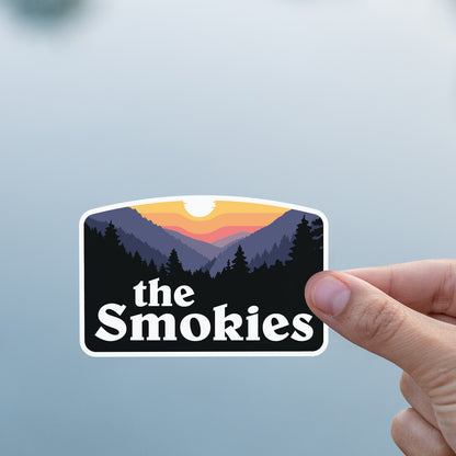 A thumb and finger pinch the The Smokies sticker against a smooth grey water surface.