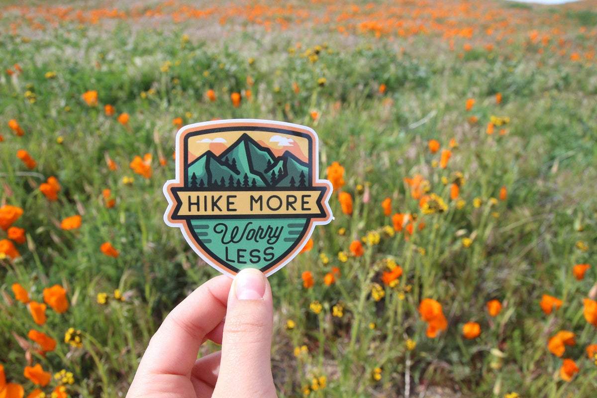 Close to a bank of orange poppies, a hand steadies the shield shaped Hike More Worry Less sticker.