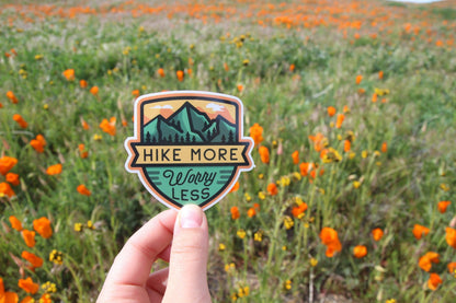 Close to a bank of orange poppies, a hand steadies the shield shaped Hike More Worry Less sticker.