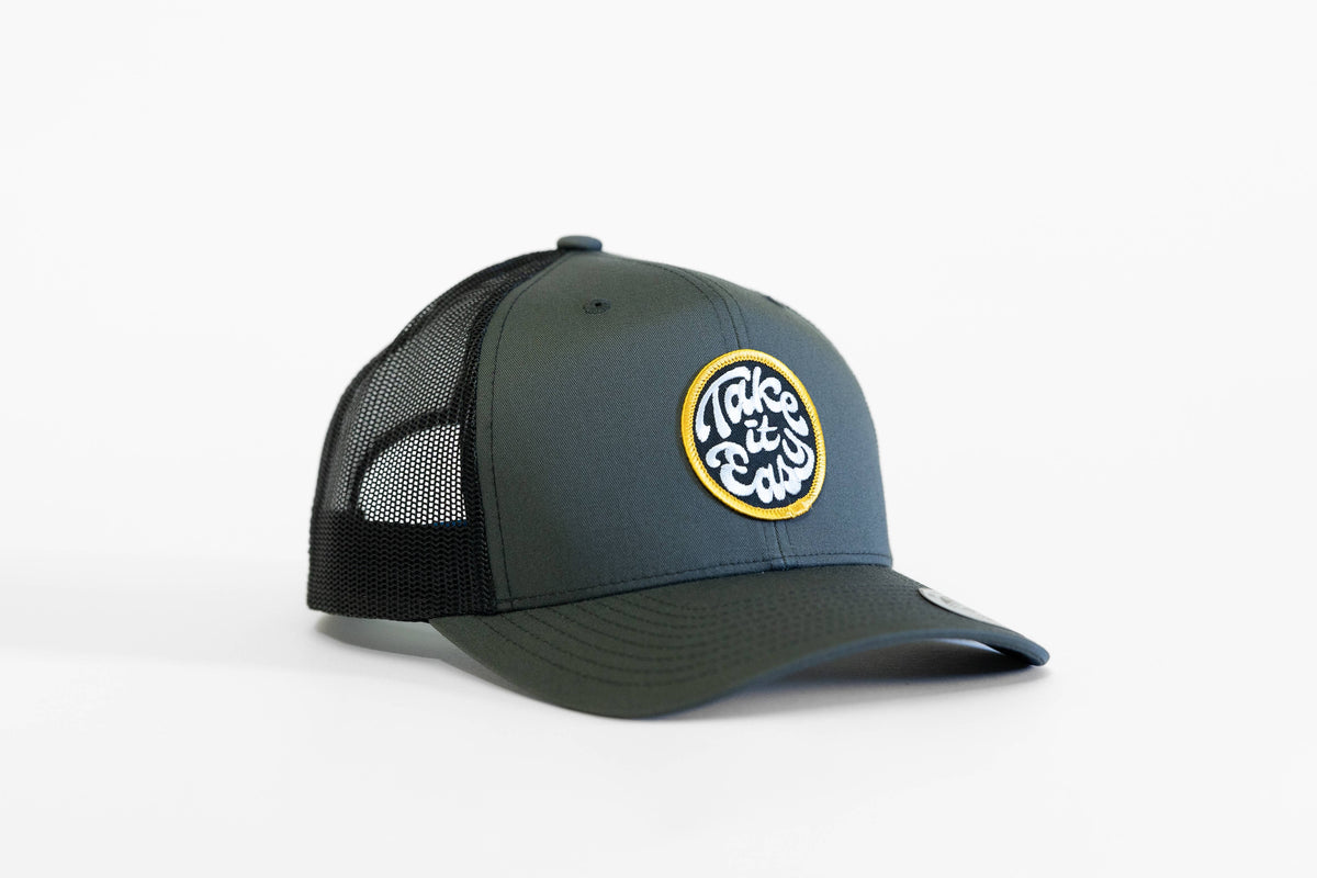 Charcoal grey Take it Easy trucker hat with black mesh sides and the circular script patch on the front.