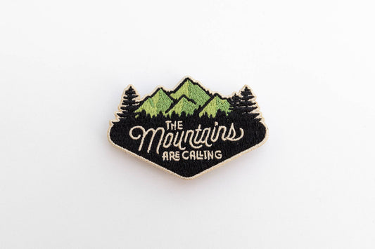 On plain white, The Mountains Are Calling patch shows green peaks, dark pines and cream script stitching.