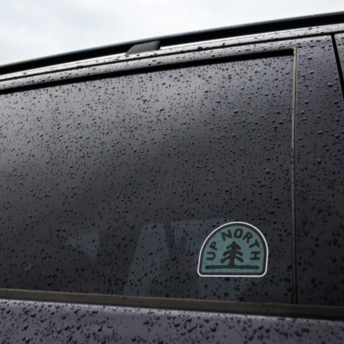 Rain beads on a dark car window where the Up North sticker rests near the lower corner.