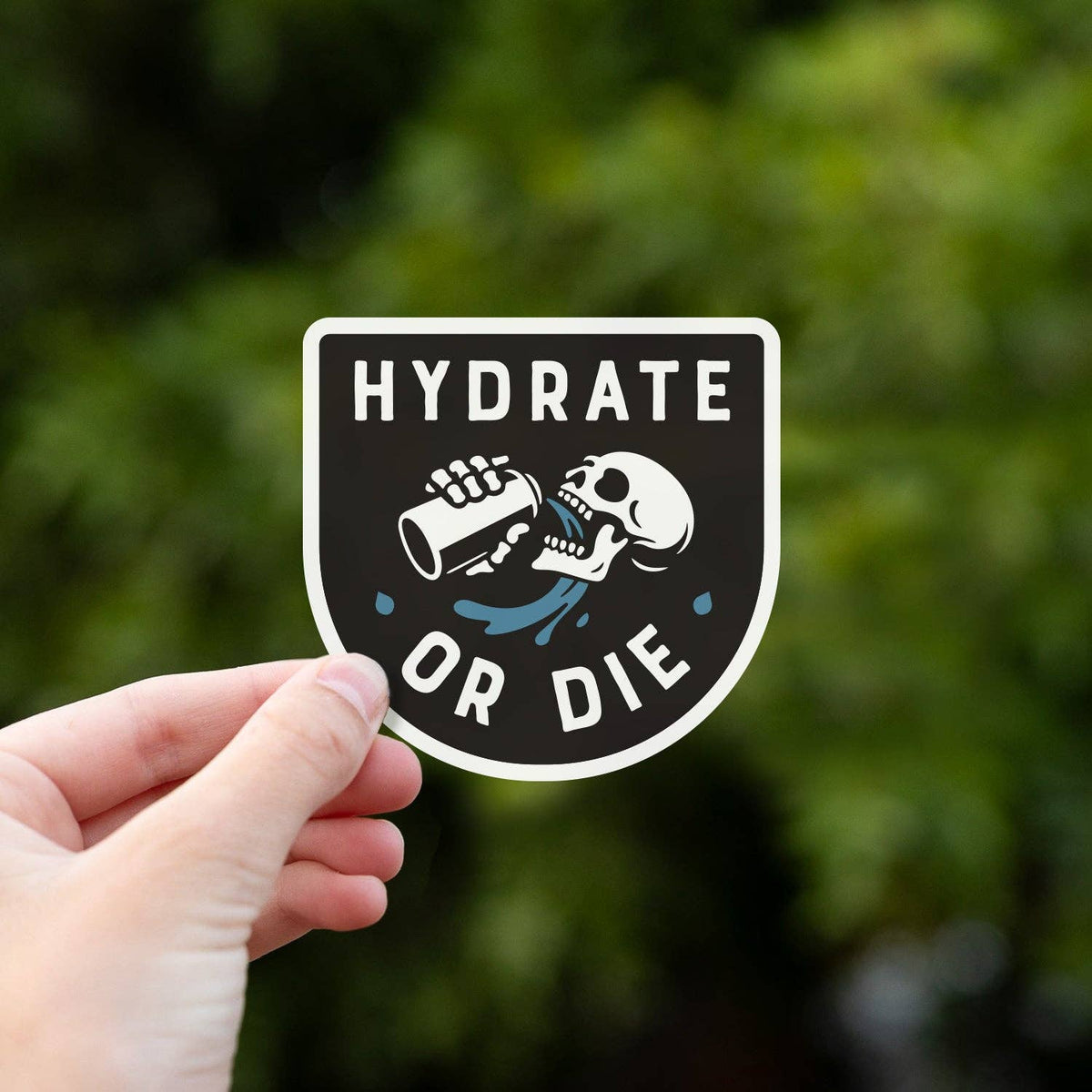 A hand holds the Hydrate or Die sticker up against a blurred wall of green leaves.