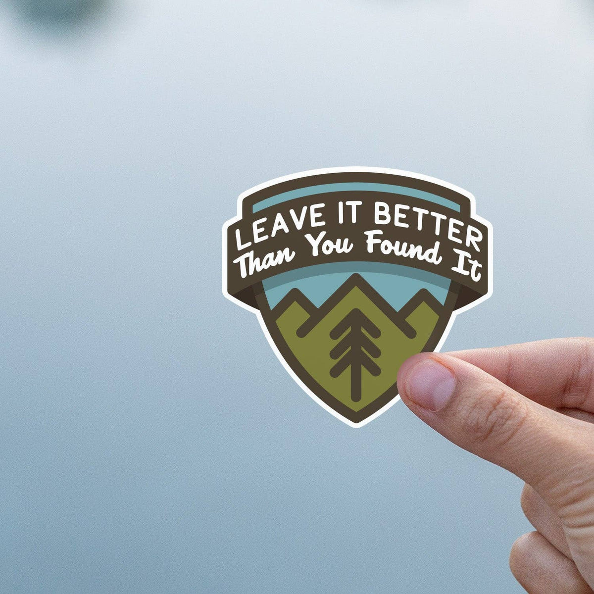 Against a soft grey blue background, a thumb steadies the Leave It Better Than You Found It sticker.