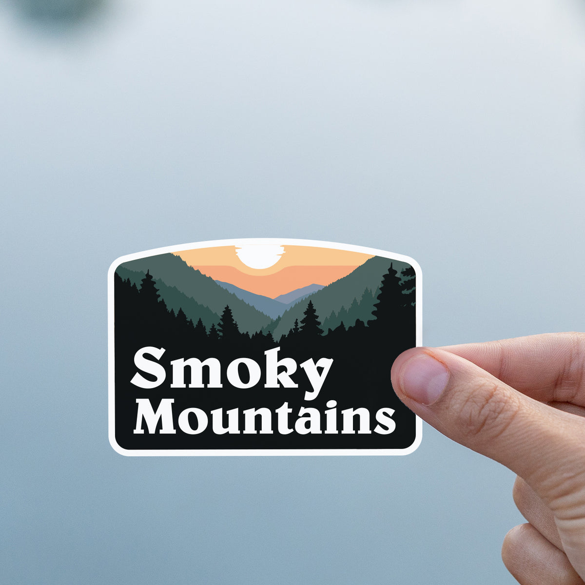 A thumb and finger hold the Smoky Mountains sticker against a calm, pale grey body of water.