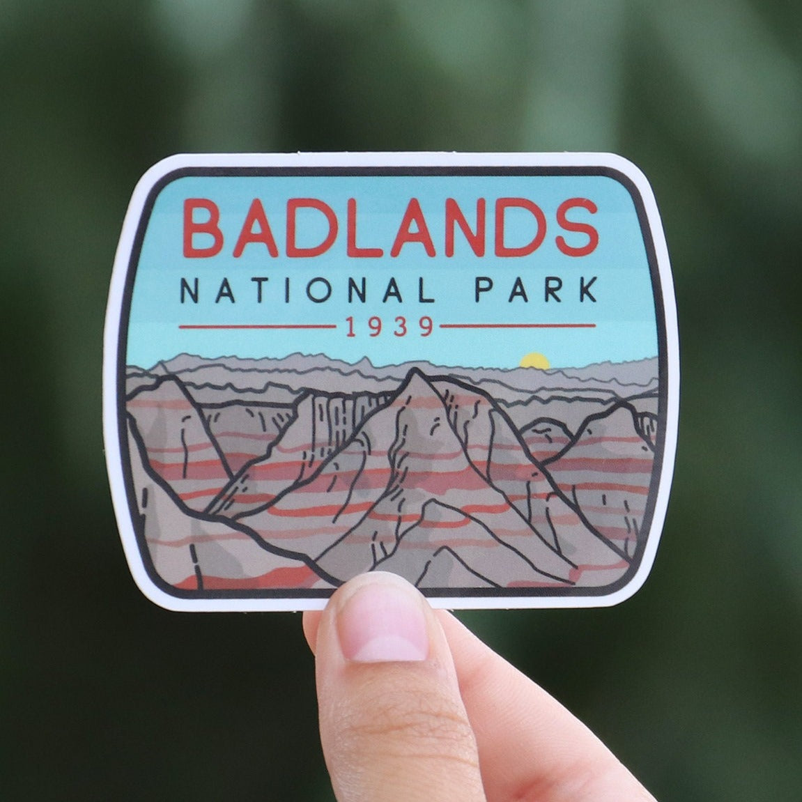 Pulled back a little, the Badlands National Park sticker sits small in a ringed hand before a pine tree.