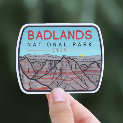Pulled back a little, the Badlands National Park sticker sits small in a ringed hand before a pine tree.