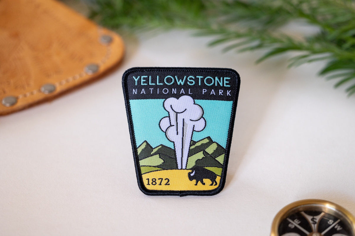 Laid on a pale surface near a leather sheath and brass compass, the Yellowstone National Park patch shows a geyser and bison.