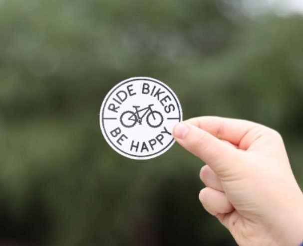 The round Ride Bikes Be Happy sticker sits pinched in a hand with soft green foliage behind it.