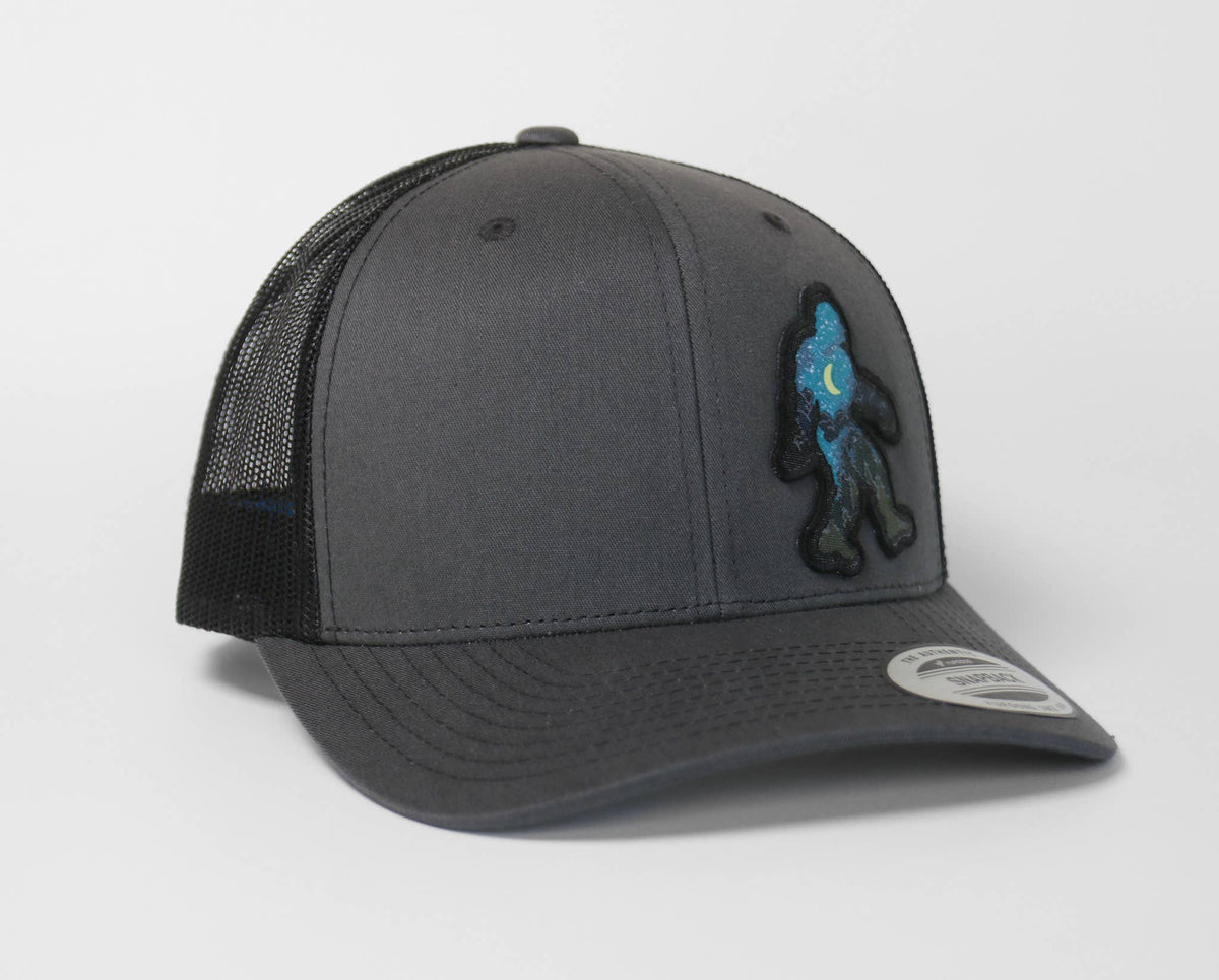 Angled left, the charcoal Bigfoot Night Sky trucker hat sets its glowing Sasquatch patch near the crown seam.