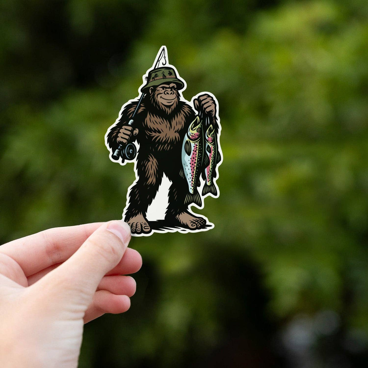 A hand lifts the Bigfoot Fishing sticker in front of thick green leaves, sunlight scattered behind.