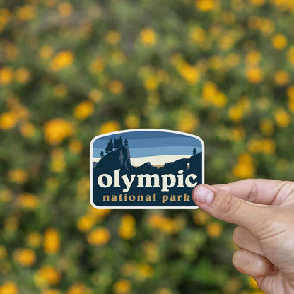 Yellow wildflowers fill the background behind the Olympic National Park sticker held at fingertip.